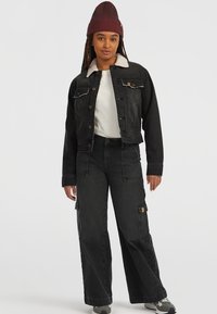 Sort denimjakke med et shearling-krave, parret med brede sorte cargo-bukser. Modellen bærer en bordeaux strikket beanie og sneakers.
