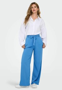 Chemise blanche surdimensionnée boutonnée, associée à un pantalon bleu à jambes larges avec un détail de taille nouée et des baskets blanches. Tissu lisse et léger.