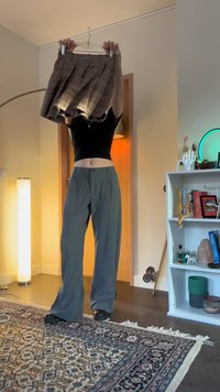 Person i svart magtröja och grå byxor håller upp en brun rutmönstrad kjol på en galge i ett mysigt rum med en lampa och bokhylla.