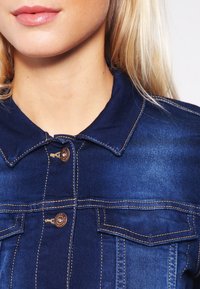 Mörkblå denimjacka med krage, guldtråd, och två bröstfickor, med bronsknappar och en mjuk yta.