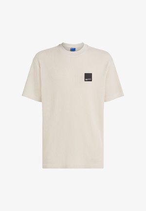 Jasnobeżowy prążkowany t-shirt z okrągłym dekoltem i krótkimi rękawami. Na lewej piersi znajduje się czarna kwadratowa metka z białym napisem.