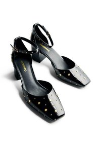 Talons en vernis noir avec un bout carré, bride de cheville et clous dorés disposés de manière uniforme sur la surface. Design de talon épais et carré.