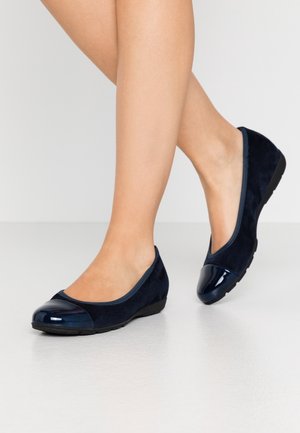 Nogi ubrane w granatowe baleriny z błyszczącymi czubkami i czarnymi podeszwami, stojące na gładkiej jasnej podłodze.