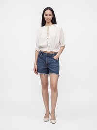 Femme debout portant un chemisier court blanc, un short en jean bleu coupé et des talons hauts blancs pointus, main dans la poche, sur fond blanc.