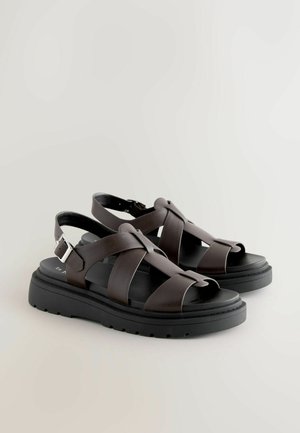 Brūnas ādas sandales ar krusteniskām siksnām, atvērtu purngalu un regulējamu potītes sprādzi. Bieza melna zole ar strukturētu virsmu.