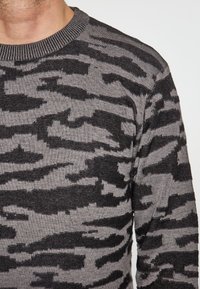 Pulover gri și negru cu un model texturat de camuflaj, având un guler rotund cu nervuri și mâneci ajustate.