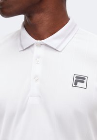Homme portant un polo blanc à col avec trois boutons et un petit logo noir et blanc sur la poitrine.
