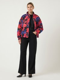 Moteris stovi vilkėdama spalvingą gėlių rašto pamuštą striukę, juodą viršų, juodas plačias kelnes ir juodus aukštakulnius batus prieš vienodą foną.