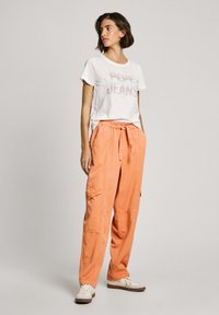 Bijela pamučna majica s printom "Pepe Jeans", uparenim s obilnim narančastim cargo hlačama koje imaju bočne džepove i pojas s vezicom.