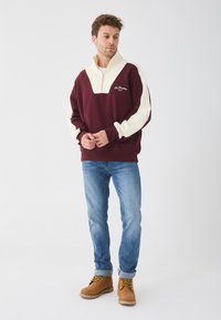 Pullover in fleece bordeaux e crema con mezza zip, caratterizzato da un design a forma di V, polsini a coste e dettagli ricamati. Indossato con jeans blu e stivali.