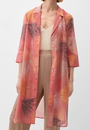 Manteau long léger corail avec des motifs abstraits de feuilles orange, blanc et noir, porté sur un haut sans manches beige et un pantalon taupe.