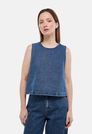 Femme à la peau claire et aux cheveux bruns raides portant un haut sans manches en denim bleu et un jean taille haute assorti, debout devant un fond blanc.