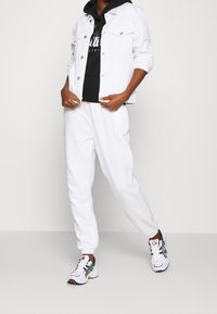 Giubbotto in denim bianco sopra una felpa nera con cappuccio, abbinato a pantaloni della tuta bianchi e scarpe da ginnastica multicolori. Vestibilità rilassata con polsini a costine.