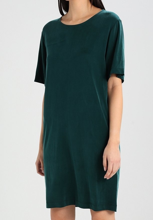 Femme portant une robe ample verte foncée, à manches courtes, longueur genou, avec un col rond, sur un fond uni.