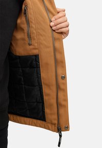 Tan jakke med et struktureret yderstof, sort kviltet foring, lynlåslomme og metalaccentuer, med en glat lynlåslukning.