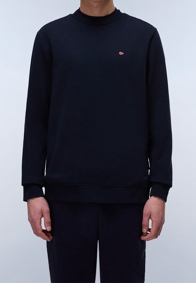 Homme portant un sweat-shirt uni bleu marine foncé à manches longues avec un petit logo rouge et blanc sur la poitrine, debout devant un fond clair.