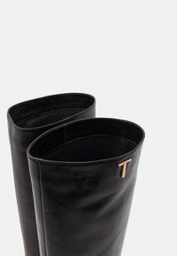 Bottes noires en cuir, montantes jusqu'au genou, avec des ouvertures ovales, une texture lisse et un accent en or sur le côté en forme de "T".