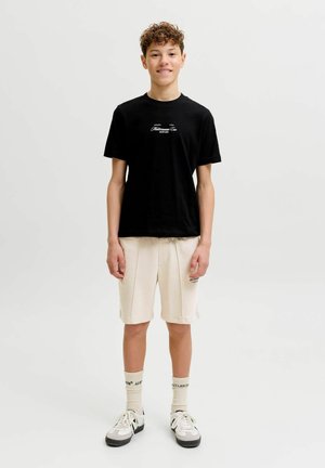 Teenager står, iført en sort t-shirt med grafik, cremefarvede shorts, hvide mellemlange sokker og hvide sneakers med sorte detaljer.