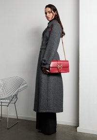 Šedý vlnený kabát s opaskom v páse, dlhými rukávmi a gombíkovými detailmi; skombinovaný s červenou koženou kabelkou s ozdobným zlatým zapínaním.