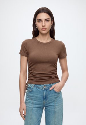 Ung kvinde med glat brunt hår iført brun ruched kortærmet top og højtaljede blå jeans, stående mod ensfarvet hvid baggrund.