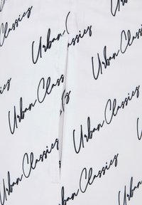 Fehér szövet, amelyen egy ismétlődő fekete kézzel írt „Urban Classics” minta látható, látható varrással és egy rövid cipzár részlettel.