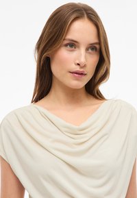 Beige drapert topp laget av myk stoff, med cowl-hals og korte ermer, med glatt tekstur og minimale designdetaljer.