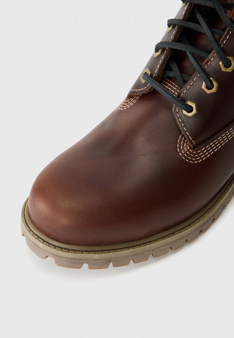 Brown leather boot with beige stitching, black laces, metal eyelets, and textured beige rubber sole on a white background.