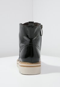 Botín de charol negro con un acabado suave, cierre de cremallera y suela beige. Presenta detalles decorativos de brogue a lo largo de los lados.