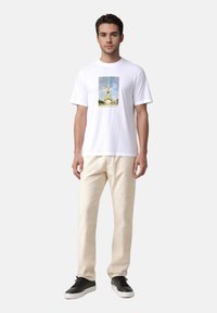 Λευκό βαμβακερό μπλουζάκι με πολύχρωμο γραφικό τύπωμα του Πύργου του Άιφελ στο μπροστινό μέρος, συνδυασμένο με μπεζ παντελόνι και μαύρα αθλητικά παπούτσια.