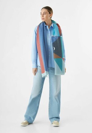Écharpe bleue à motifs portée sur une chemise bleu clair ; présente des formes géométriques dans des nuances de bleu, rouge et sarcelle avec des bords à franges.