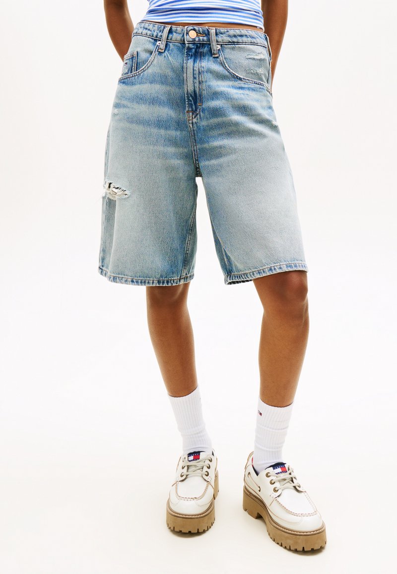 Άτομο που φοράει ανοιχτό μπλε τζιν σορτς μέχρι το γόνατο με μικρό σκίσιμο στο αριστερό μηρό, λευκές κάλτσες και λευκά παπούτσια με χοντρή σόλα.