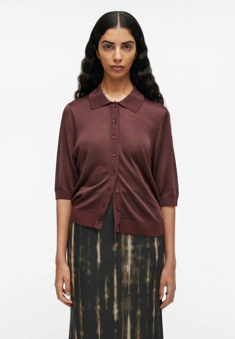 Donna con i capelli lunghi e scuri, che indossa una camicia a maniche corte color maroon con bottoni e una gonna nera con un motivo a strisce beige in techniche tie-dye, in piedi su uno sfondo bianco.