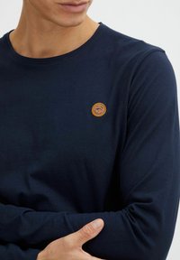 Navy long-sleeve shirt made of soft cotton, featuring a round neckline and a small round brown leather logo patch on the chest.