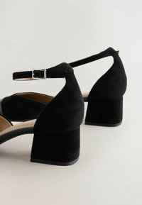Sandales à talon bloc en daim noir avec une bride à la cheville, côtés ouverts et doublure intérieure lisse. Caractérisées par un bout carré et un talon robuste.