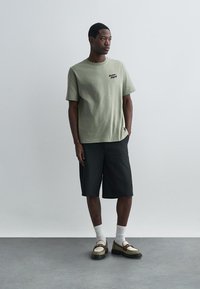 Olijfgroen t-shirt met korte mouwen, zwarte shorts, witte sokken en meerkleurige sneakers van een gestructureerde stof met minimale merkdetails.