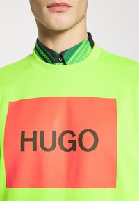 Felpa verde neon con una prominente grafica rettangolare rossa con la scritta nera "HUGO" centrata. Collo a costine e dettagli che richiamano una maglietta sottostante.