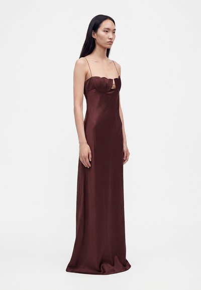 Femme portant une longue robe de soirée en satin brun foncé avec de fines bretelles et un détail de buste structuré à bords festonnés sur un fond blanc uni.