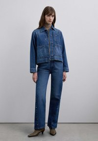 Chaqueta vaquera con detalles de botones, combinada con vaqueros de pierna recta que presentan acentos de color a lo largo de las costuras, ambos en azul oscuro. Botas marrones de tacón.