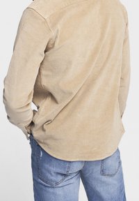 Chemise en velours côtelé beige avec une surface texturée, fermeture à boutons et un ourlet arrondi, associée à un jean en denim bleu clair avec poches arrière.