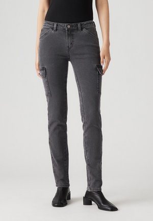 Personne portant un pantalon cargo gris foncé slim avec des poches à rabat sur les cuisses et des bottines noires à talons, debout devant un fond uni.