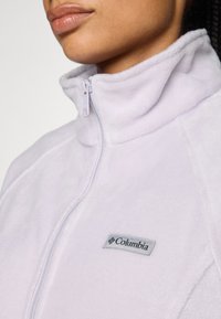 Hele lilla fliisjakki kõrge kraega ja esiärkega. Omab hõbedast Columbia logo tähistust. Pehme tekstuur, päevase disain.
