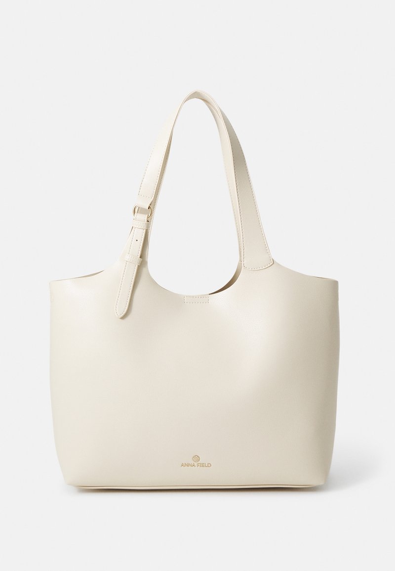 Sac fourre-tout en cuir couleur crème avec bandoulière ajustable et petit logo doré "ANNA FIELD" au centre devant.
