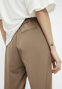 Pantalons marron avec une taille élastique, dotés d'une seule poche arrière et d'une texture lisse. Le modèle porte un haut blanc ample.