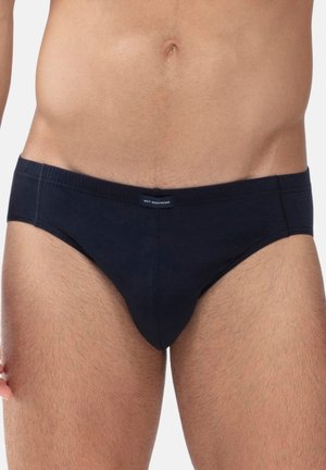 Männlicher Torso trägt schwarze Slip-Unterwäsche, zeigt Taille, unteren Bauch und obere Oberschenkel vor einfachem Hintergrund.
