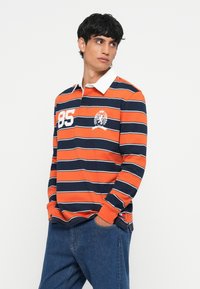 Camisa polo de manga longa às riscas laranja e azul-marinho com colarinho branco, apresentando um detalhe de logótipo e o número "85" na parte da frente.