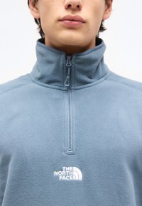 Άτομο που φοράει ανοιχτομπλέ φλις πουλόβερ με φερμουάρ μέχρι το στήθος και το λογότυπο The North Face κεντημένο στο στήθος.