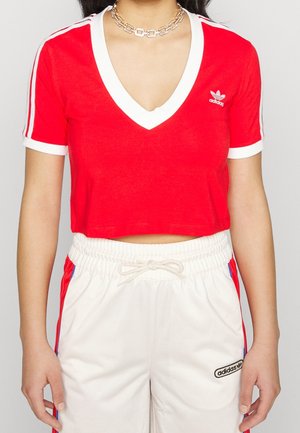 Camiseta recortada roja con escote en V, ribete blanco y mangas cortas, con rayas blancas en las mangas y un logo de Adidas en el pecho.