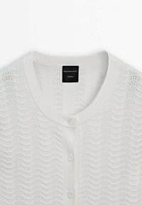 Valkoinen neulottu neuletakki, jossa on aalto pattern, napit ja ribbineck. Tunnuksessa lukee "MASSIMO DUTTI STUDIO."