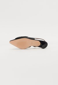 Chaussure en cuir verni noir avec un bout pointu, un talon plat, et un nœud décoratif sur le dessus. La semelle est beige avec un motif de grip texturé.