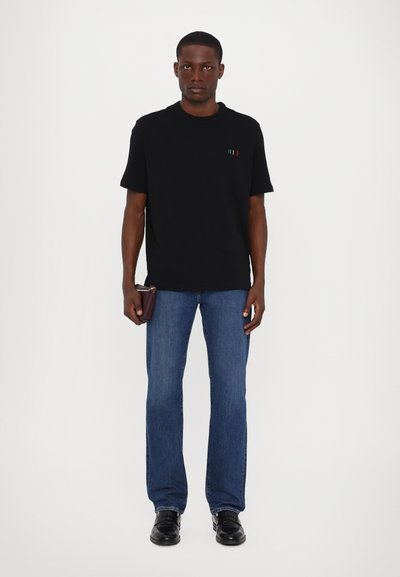 T-shirt noir à manches courtes avec des accents de rayures colorées, associé à un jean bleu à jambe droite et des chaussures noires, tenant un portefeuille à la main.